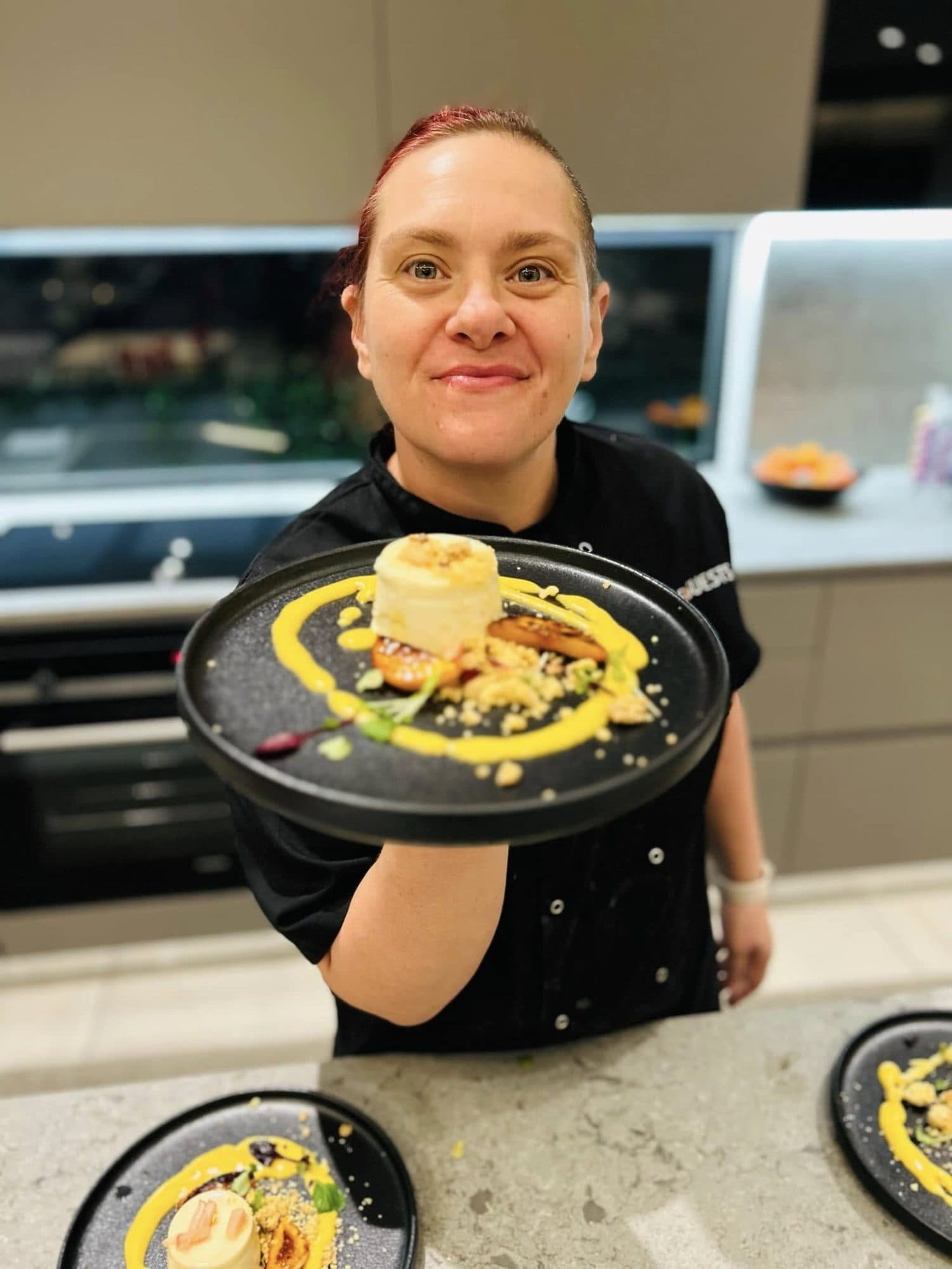 Chef Maradine smiling and presenting a plated dessert in a kitchen